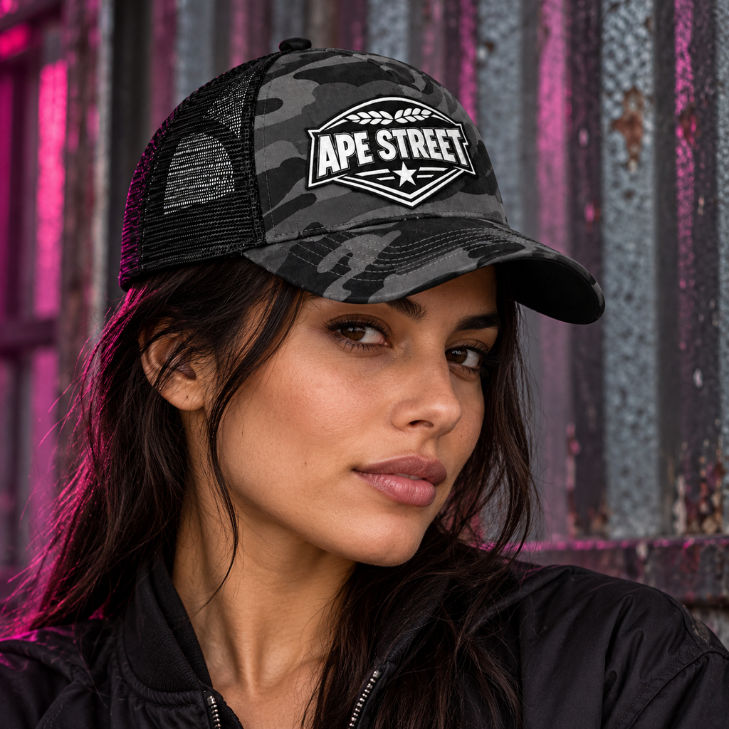 Model wearing the Ape Street black camo trucker cap against an urban metal background