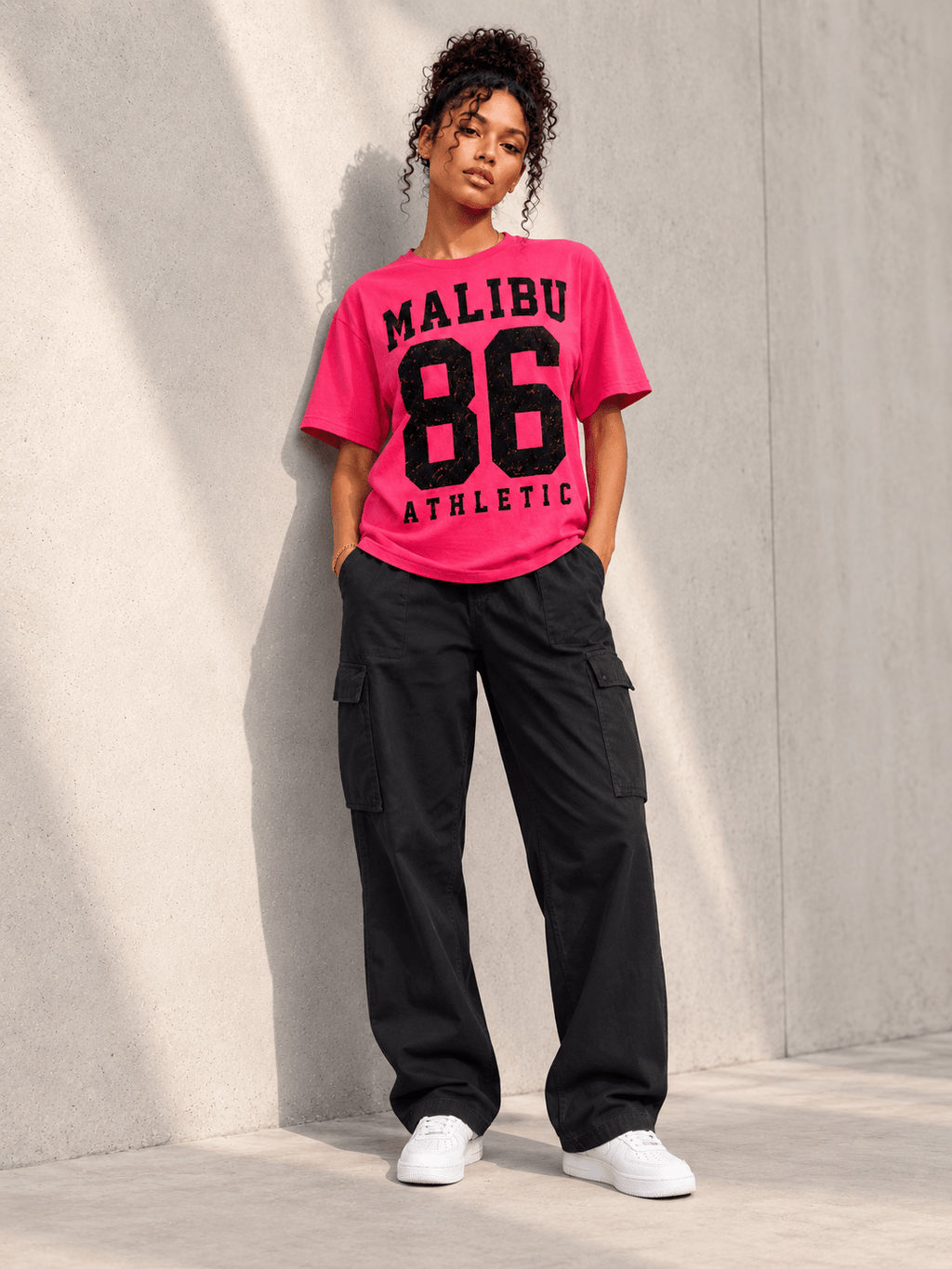 Model wearing Moda Originals Malibu 86 Tee — Pink, L, front view showing the design, against a light concrete backdrop.