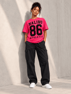 Model wearing Moda Originals Malibu 86 Tee — Pink, L, front view showing the design, against a light concrete backdrop.