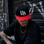 Model wearing Urban Avenue black and red UA cap against a shutter.