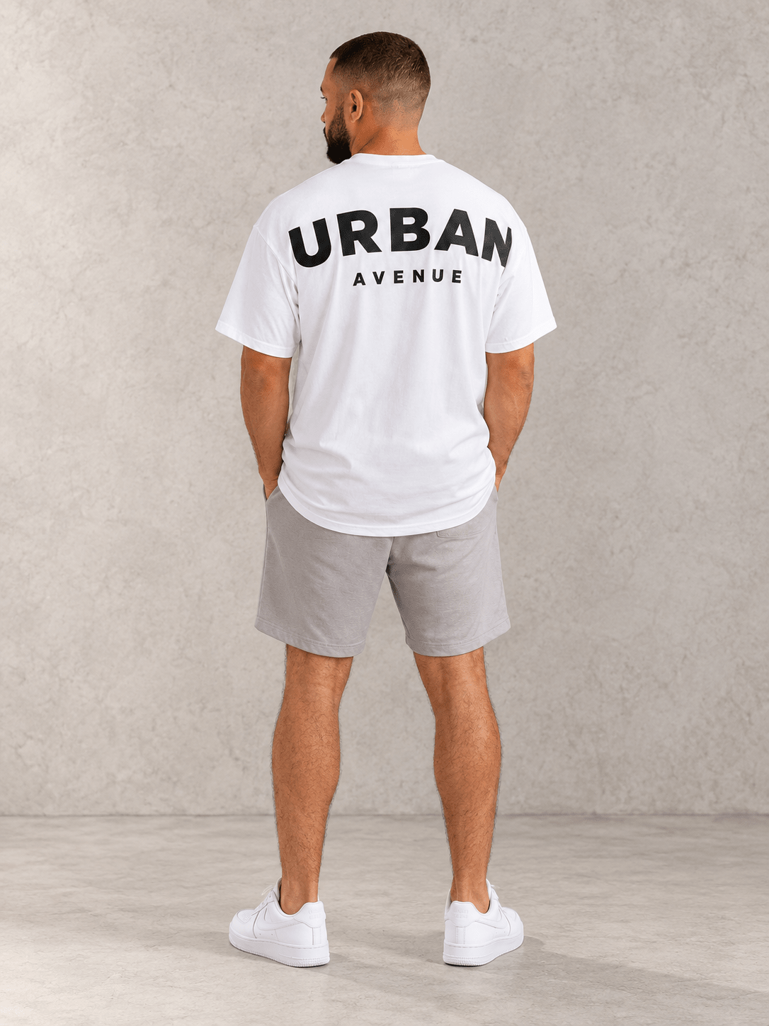 Model wearing Urban Avenue Shoulder Print Tee — White, back view showing the back print, against a light concrete backdrop.