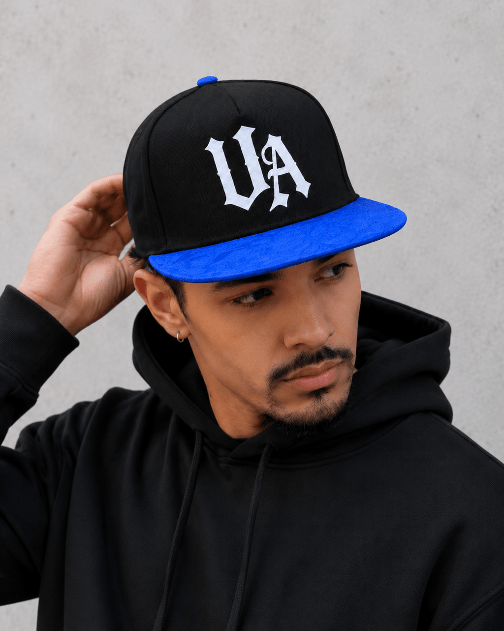 Model wearing Urban Avenue UA Logo Snapback in black and blue against a light grey catalogue background.