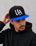 Model wearing Urban Avenue UA Logo Snapback in black and blue against a light grey catalogue background.