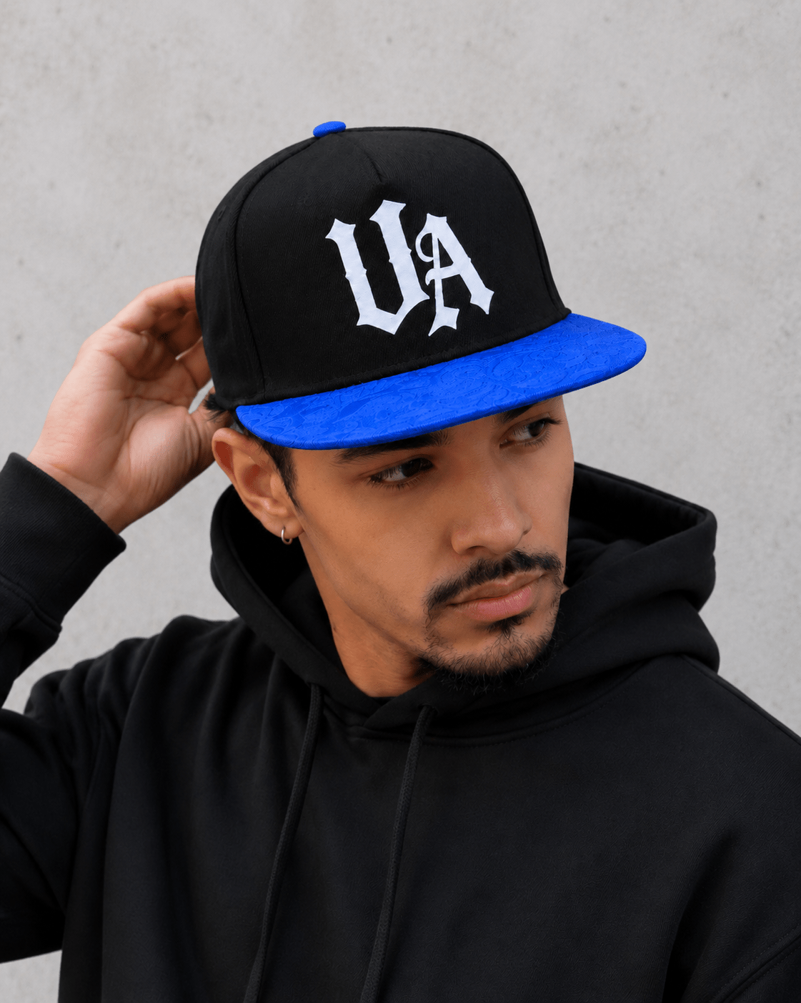 Model wearing Urban Avenue UA Logo Snapback in black and blue against a light grey catalogue background.