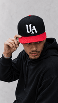 Model wearing Urban Avenue UA Logo Snapback in black and red against a light grey catalogue background.