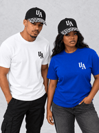 Two models wearing white and royal blue Essentials tees, clearly showing UA logos and patterned cap peaks; one cap included per pack