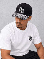 White Essentials tee model showing the Gothic UA cap logo and full black-and-white patterned peak