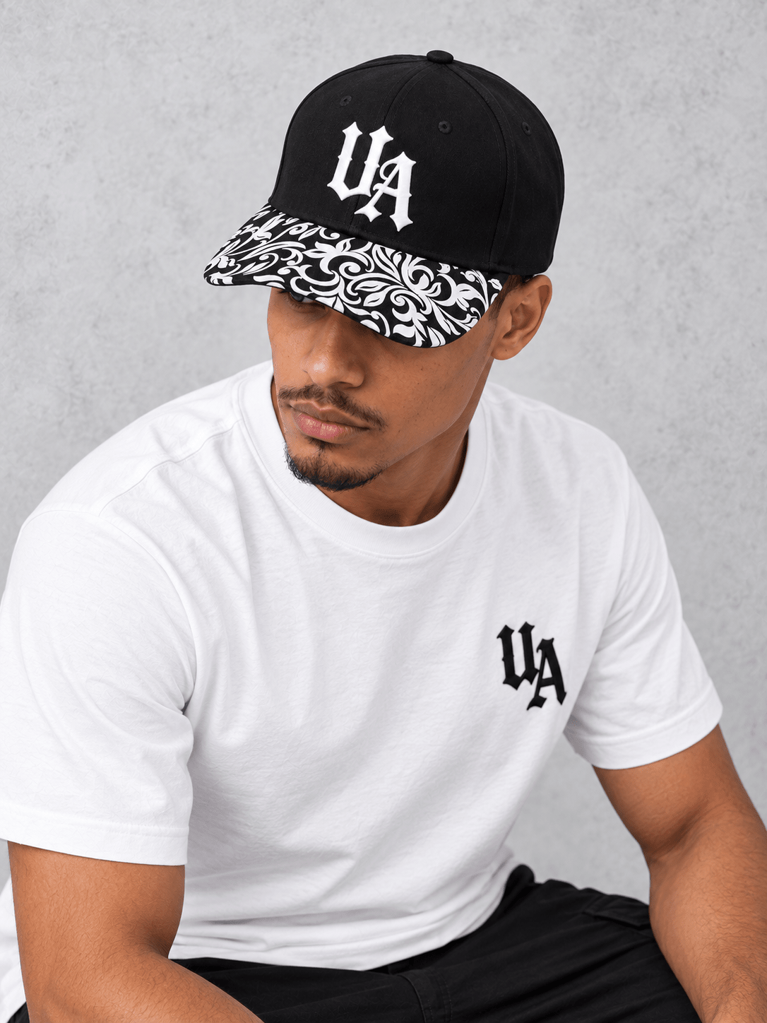 White Essentials tee model showing the Gothic UA cap logo and full black-and-white patterned peak